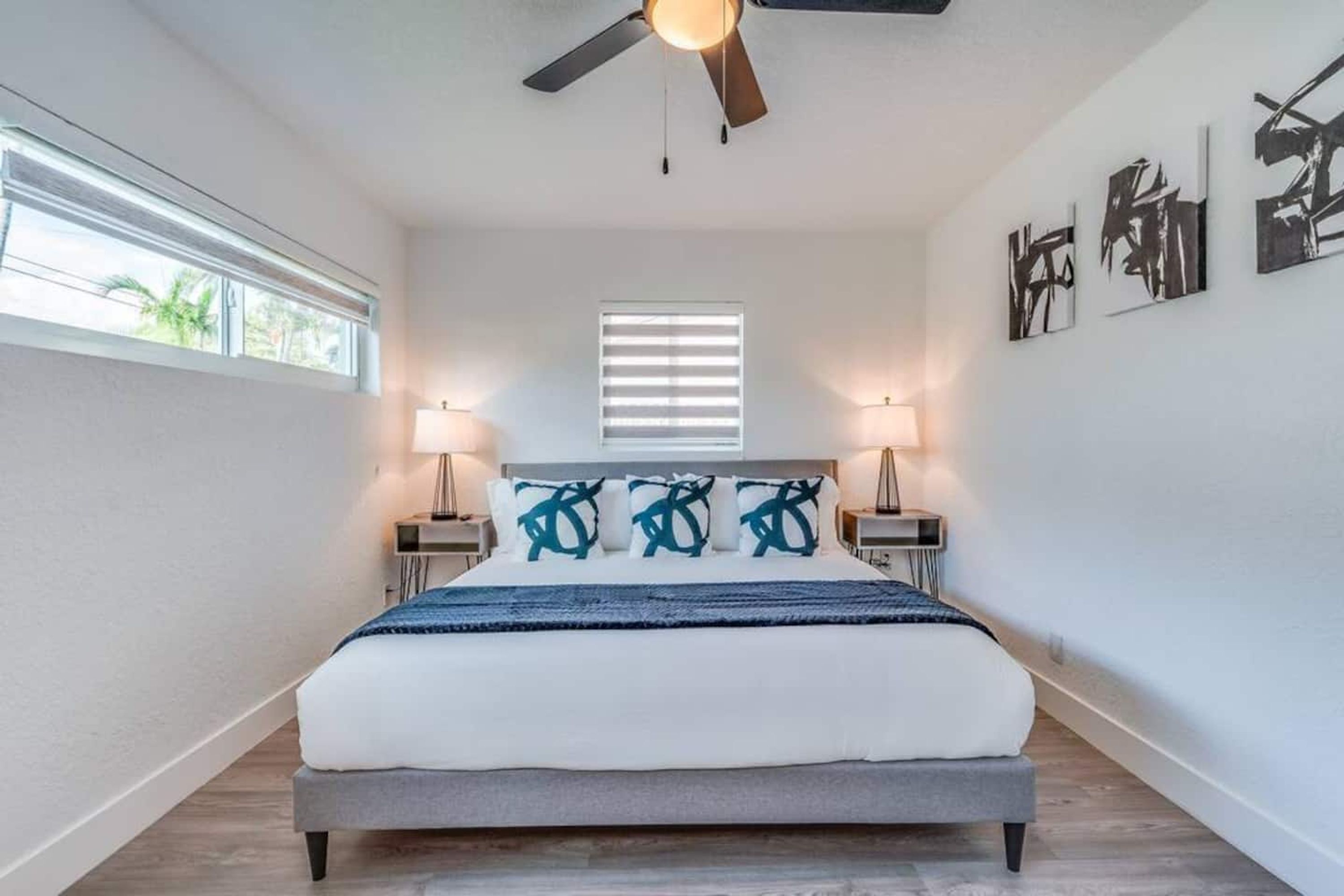 A bedroom with a bed, ceiling fan, bedside lamps, a window, and artwork on the wall.