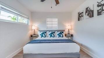 A bedroom with a bed, ceiling fan, bedside lamps, artwork on the wall, and a window.