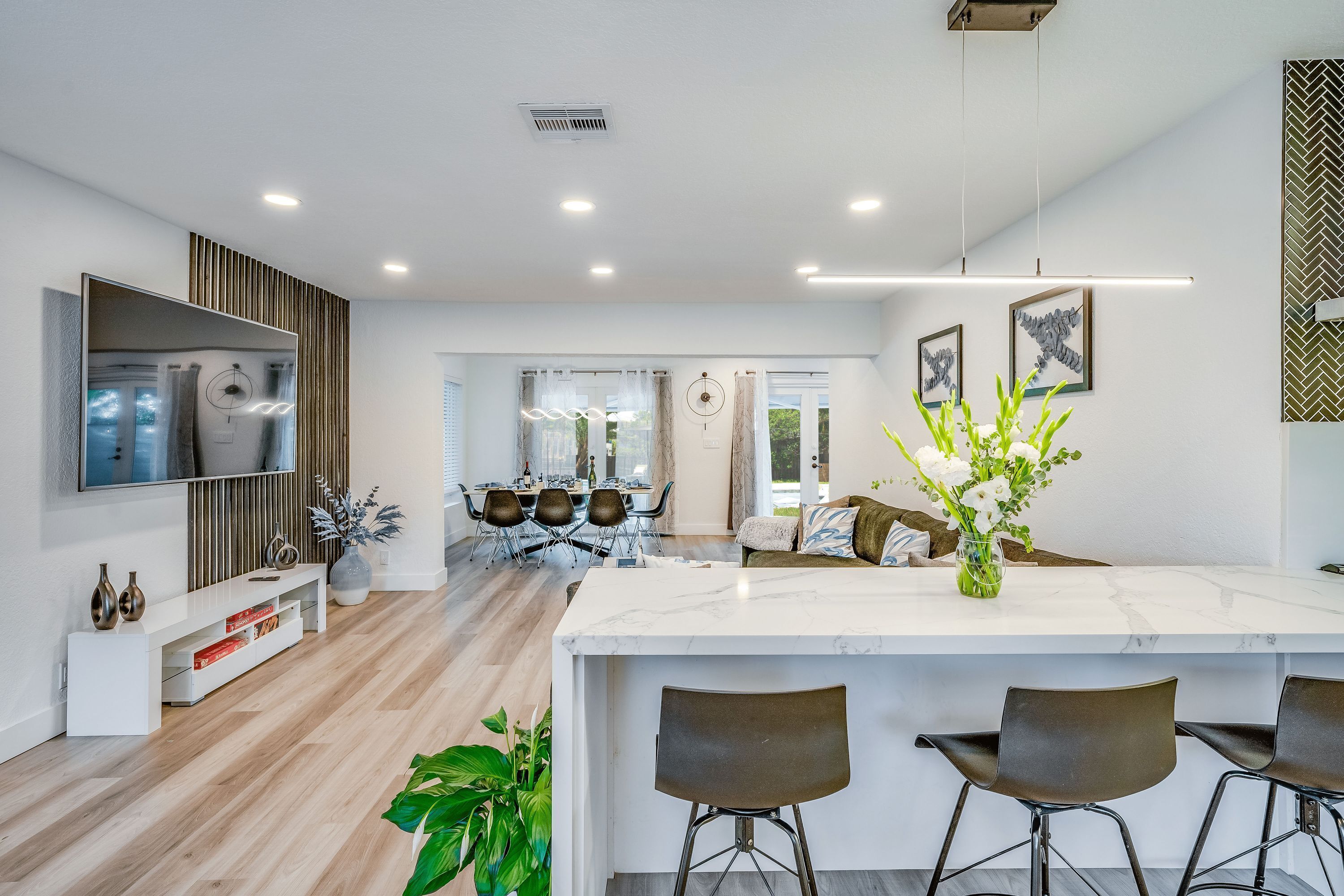 Una cocina moderna con una isla de mármol, taburetes de bar, una zona de comedor con sillas y un televisor montado en la pared.
