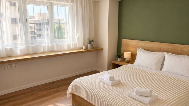 Ett hotellrum med en säng i trä, ett fönster med tunna, genomskinliga gardiner, ett nattduksbord med en lampa och en grön accentvägg.