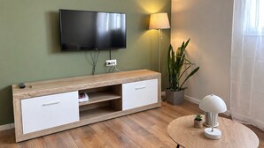 Ett modernt vardagsrum med en platt-TV, en träkonsol, vita lådfronter, ett runt bord med en lampa och en krukväxt.