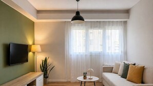 Ett modernt vardagsrum med en platt-TV, en träkonsol, ett runt soffbord, en beige soffa och ett stort fönster med tunna, genomskinliga gardiner.