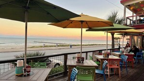 Eine Holzterrasse mit Tischen und Stühlen, die auf einen Strand und das Meer blickt.