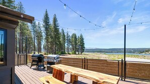 Terraza exterior con bancos de madera y una parrilla de barbacoa, con vistas a un lago y un bosque.