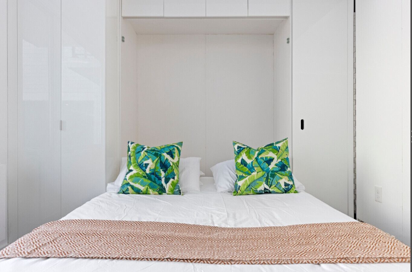 A bedroom with a bed, two pillows with a leaf pattern, a patterned throw blanket, and white walls.