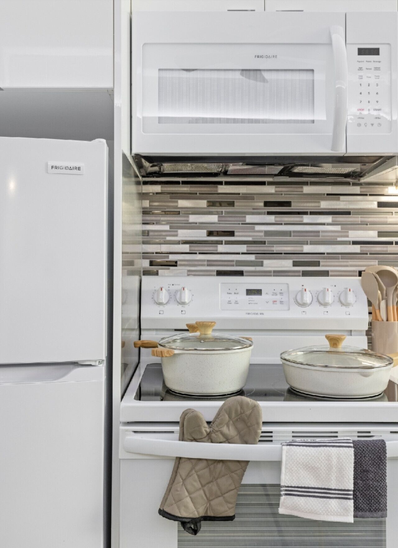 Une cuisine avec un réfrigérateur blanc, un four à micro-ondes, une cuisinière avec deux casseroles, des maniques et un porte-serviettes.