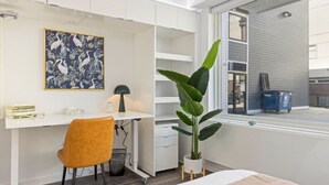 Et moderne værelse med et hvidt skrivebord, en stor plante, en seng med mønstret sengetøj og et vindue med udsigt.