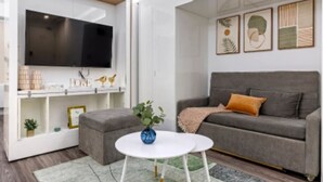 En moderne stue med en grå sofa, et hvidt sofabord, et fladskærms-tv og dekorative genstande på hylder.