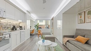 Et moderne køkken med hvide skabe, et spiseområde med et rundt bord og stole, samt en stue med sofa og sofabord.