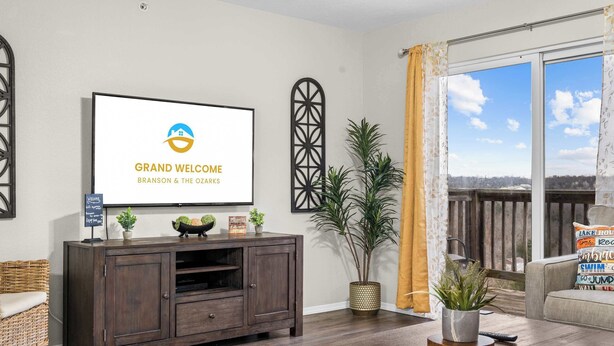 Ein Wohnzimmer mit einem Fernseher, der 'GRAND WELCOME BRANSON & THE OZARKS' zeigt, einer dunklen Holz-Unterhaltungseinheit, einem Korbstuhl, einer Couch mit Kissen und einem Blumentopf, sowie einem Blick durch ein Fenster.