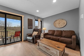 A living room with a brown leather sofa, a leather armchair, a wooden coffee table, and a view through a glass door to an outdoor area. - Hole-in-One Hideaway  For 4 (Branson)