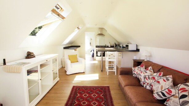 Un espacio habitable en el ático con cocina, muebles blancos, alfombra roja estampada y sofá marrón con cojines decorados.