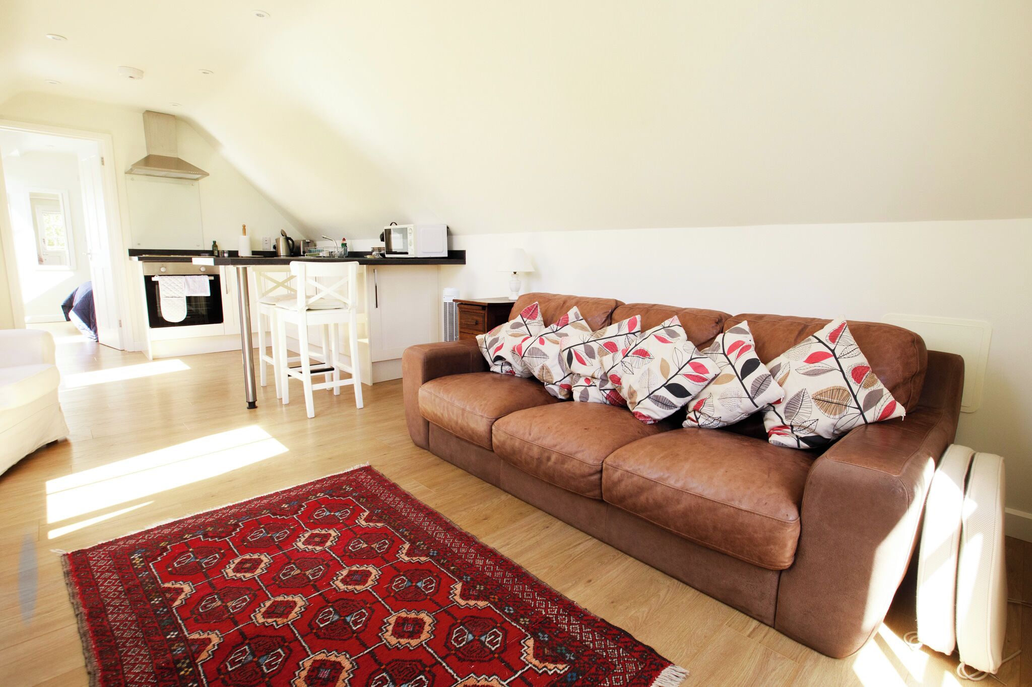 A living room with a brown sofa, patterned pillows, a red rug, a kitchen area with a microwave, and a white chair.