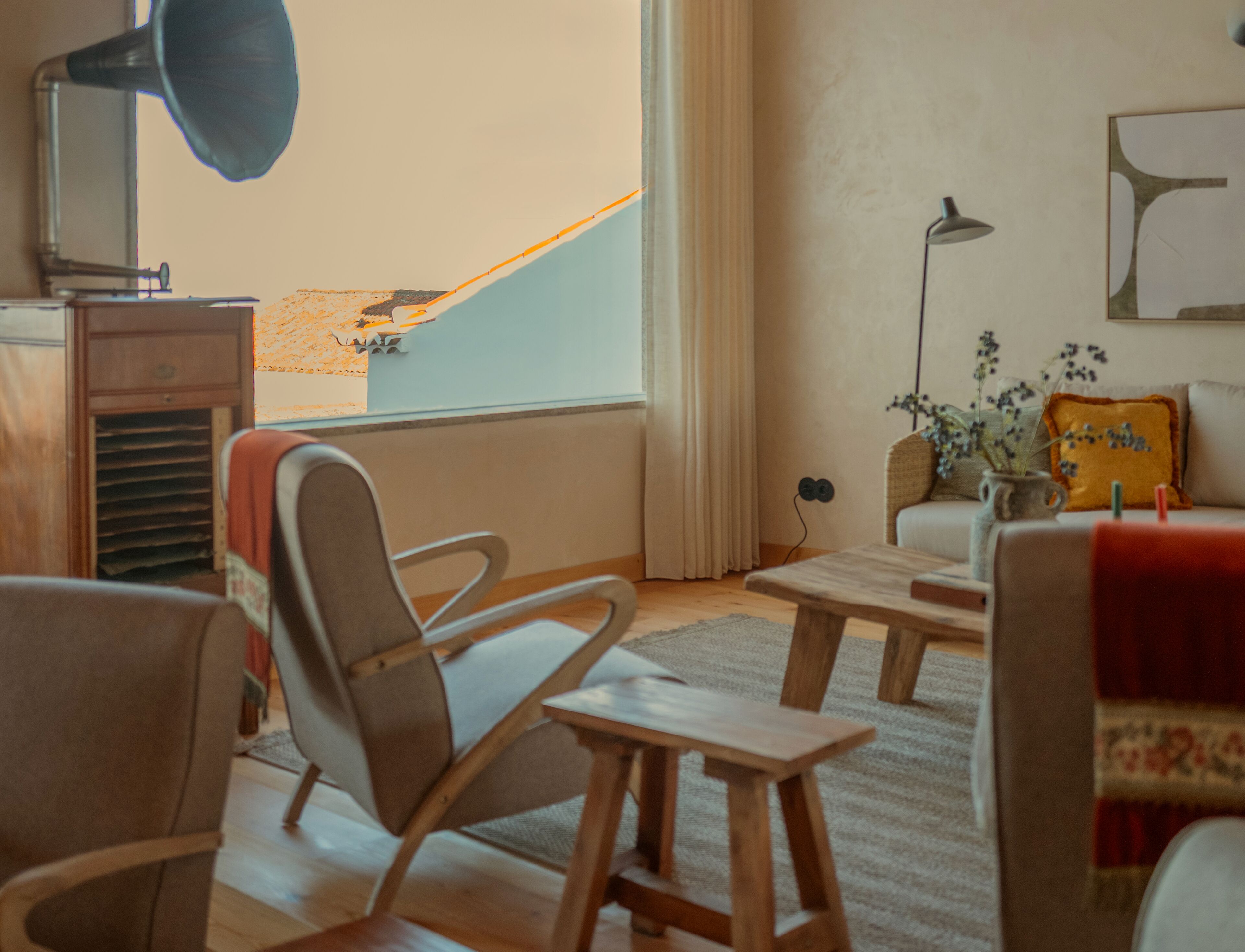 A room with a wooden chair, a small table, a sofa, a lamp, and a view of a building outside.