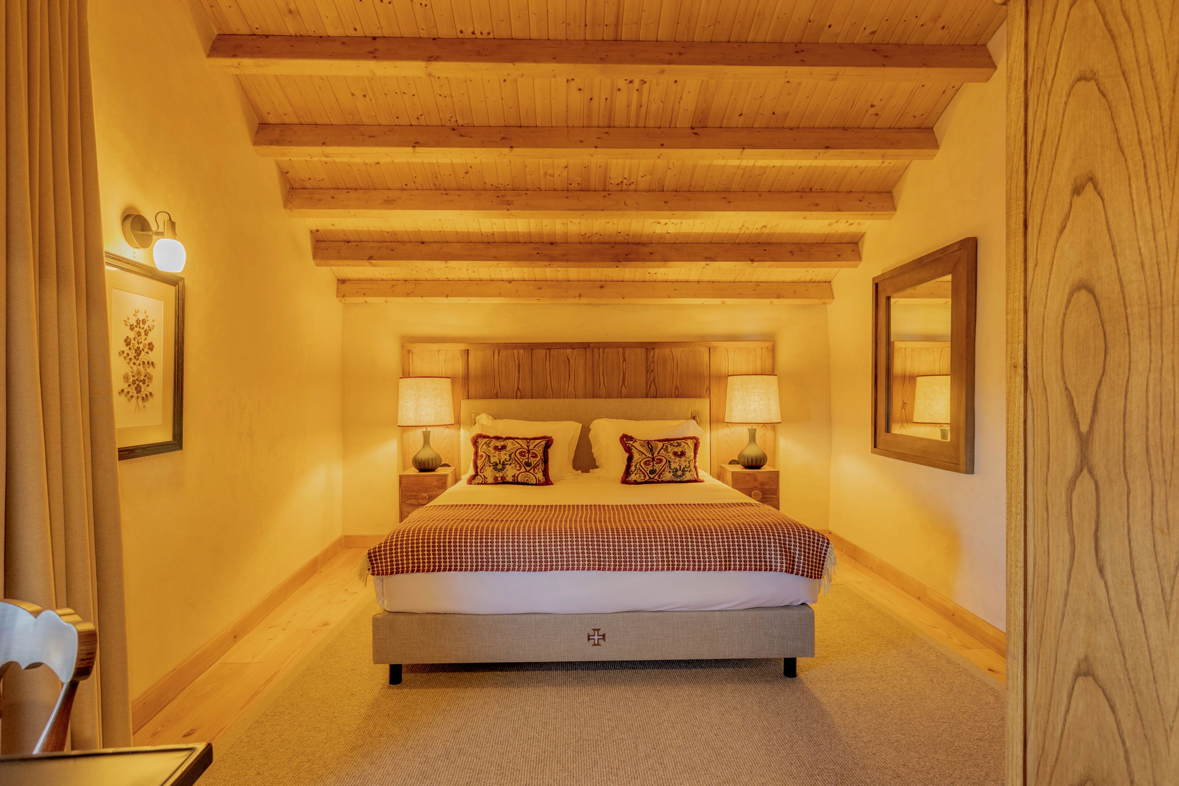 Ein Schlafzimmer mit Holzboden, einem Bett mit kariertem Überwurf, zwei Nachttischlampen und einem gerahmten Bild an der Wand.