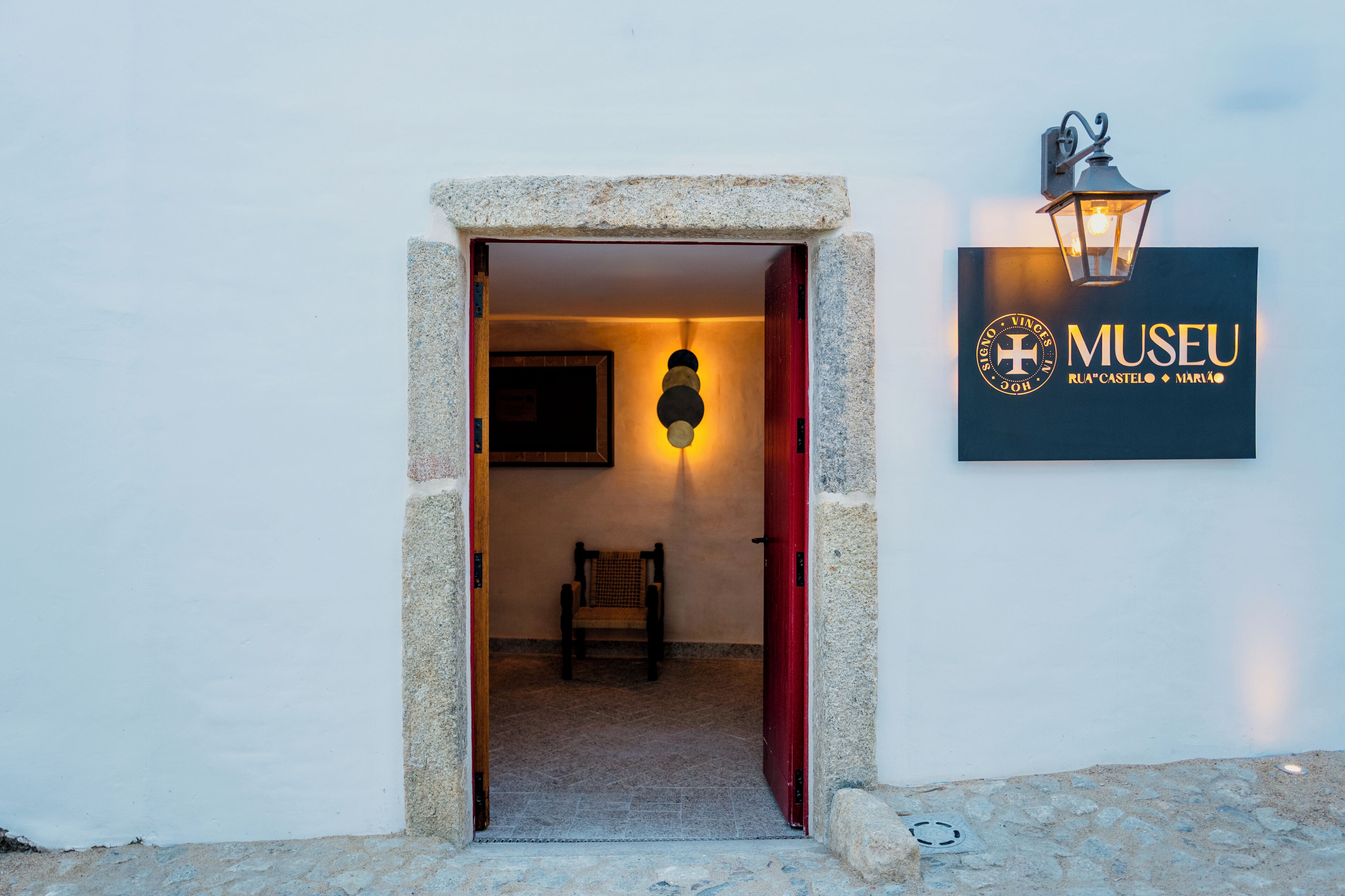 An entrance to a building with a stone archway, a chair inside, and a wall-mounted sign with the word 'MUSEU' and a logo.