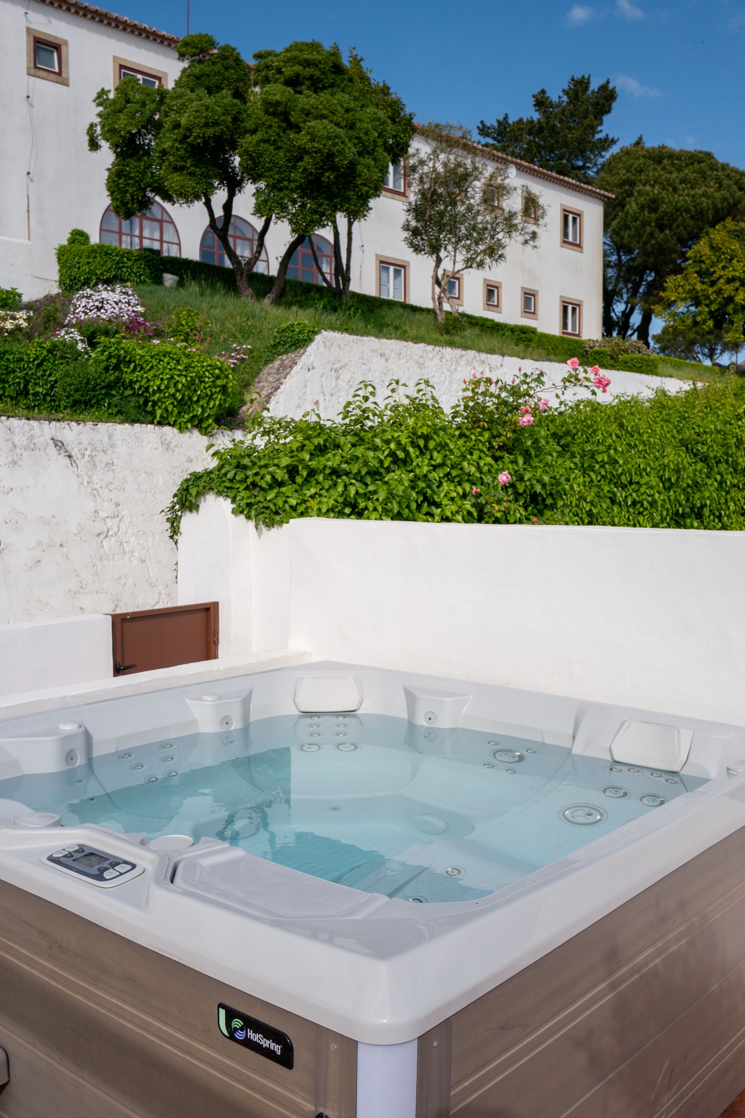 A hot tub with a control panel, surrounded by lush greenery and a white building with arched windows in the background.