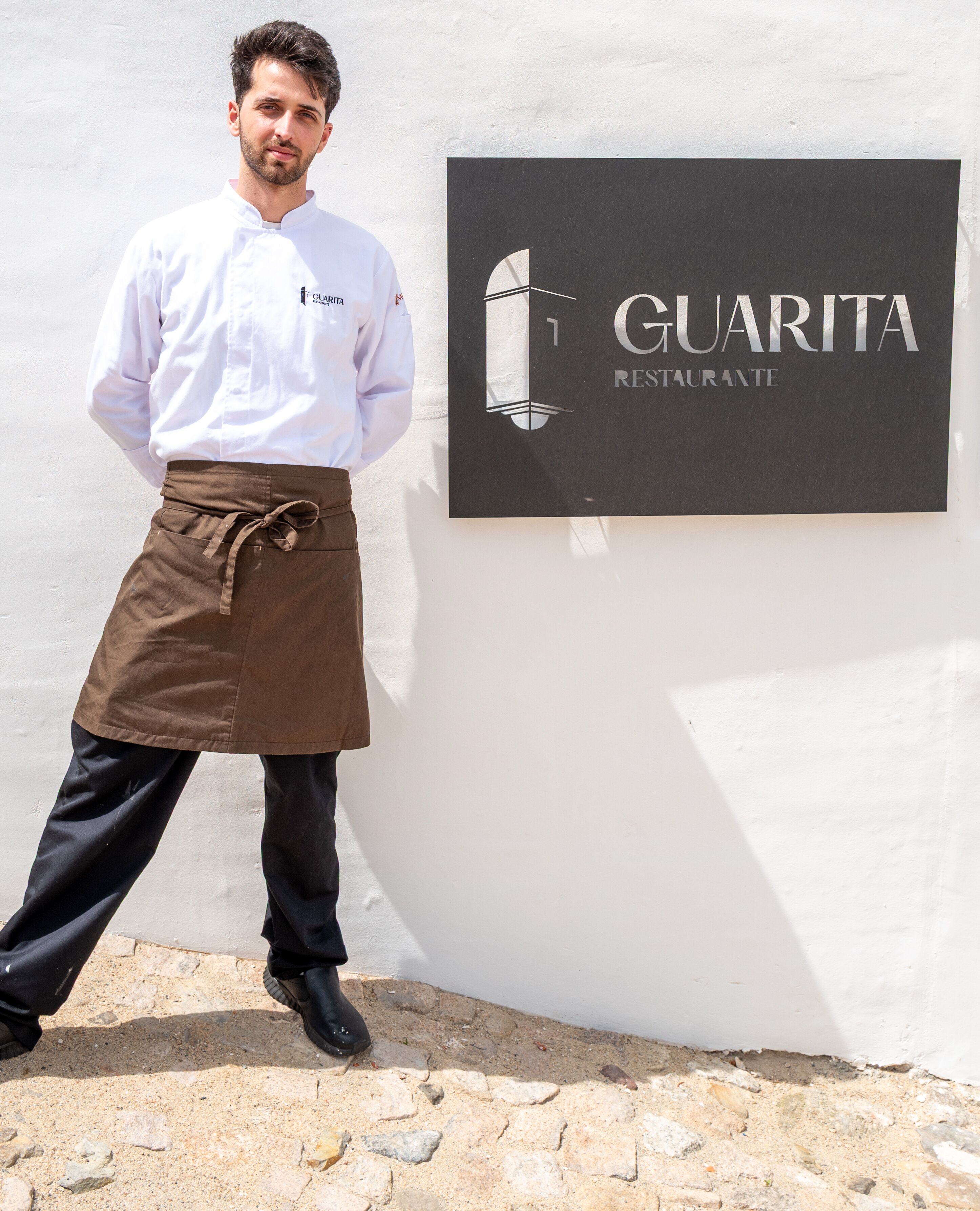 A person in chef attire standing next to a sign that reads 'GUARITA RESTAURANTE'.