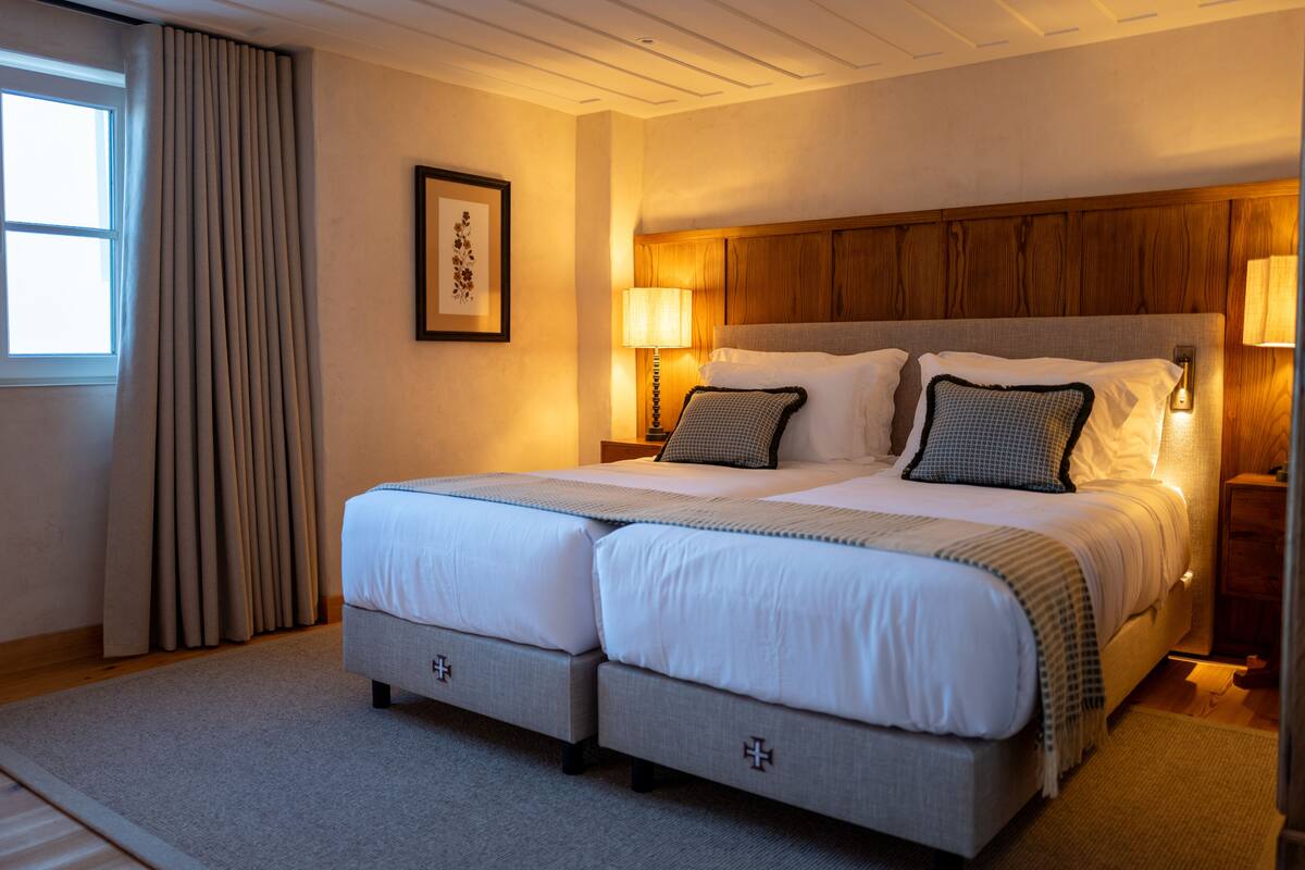 A double bed with a wooden headboard, two bedside tables with lamps, a framed picture on the wall, and a window with curtains.