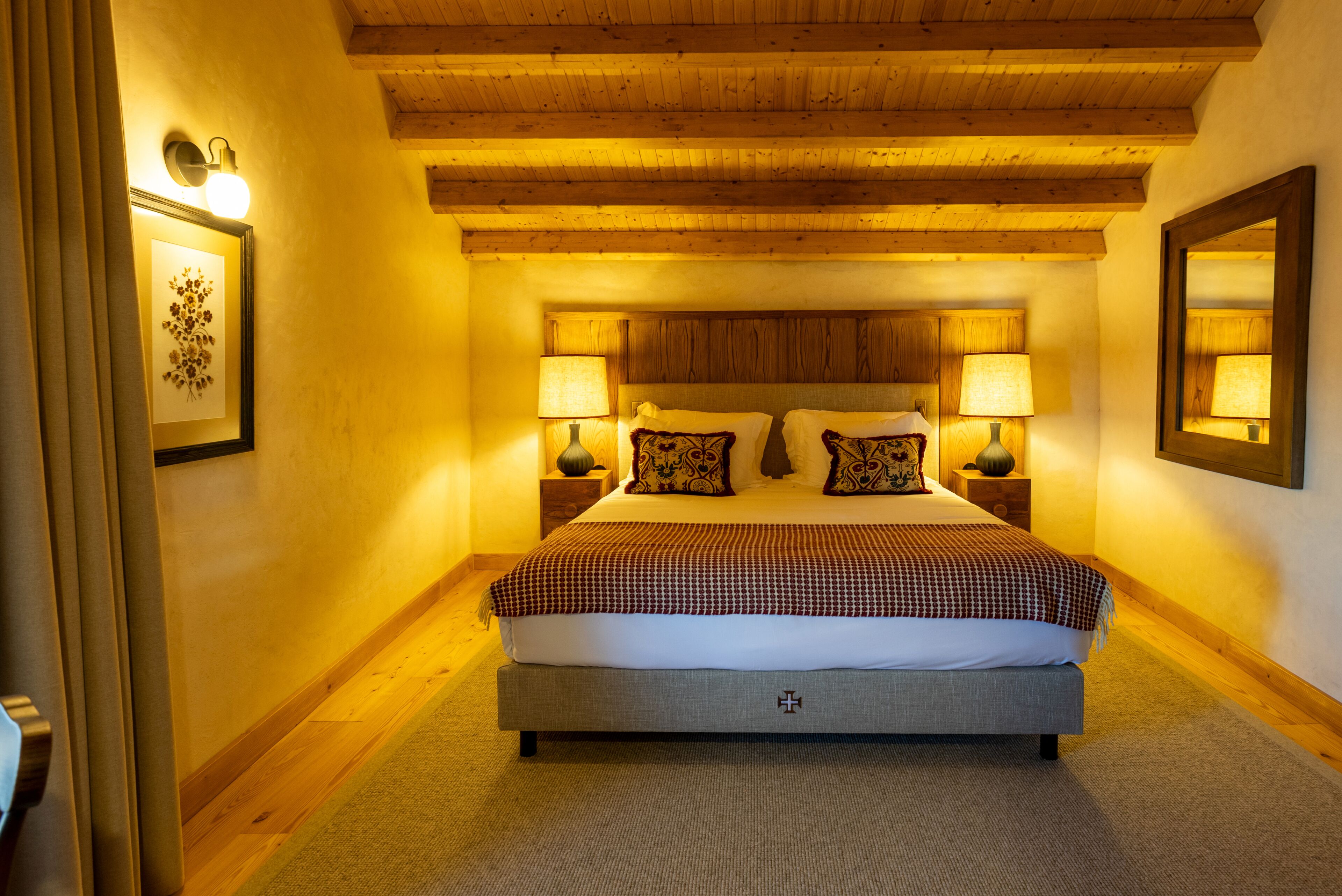 Un dormitorio con una cama grande, dos mesitas de noche con lámparas, vigas de madera en el techo y un cuadro enmarcado en la pared.