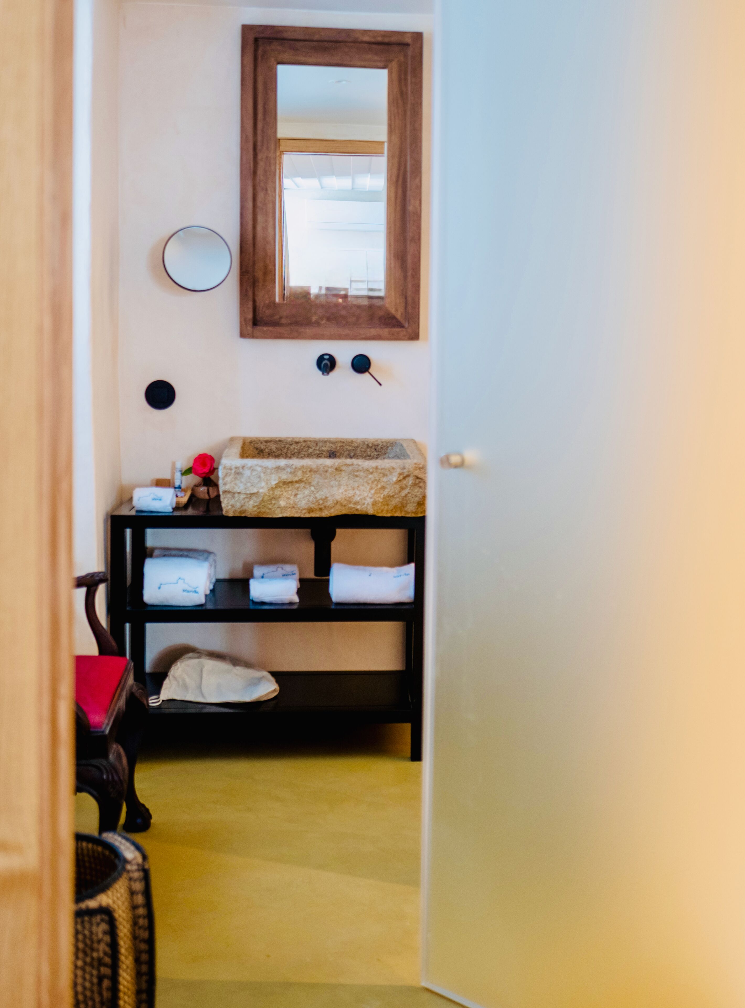 A narrow hallway with a wooden door, a mirror, a shelf with folded towels, and a small table with a red object.