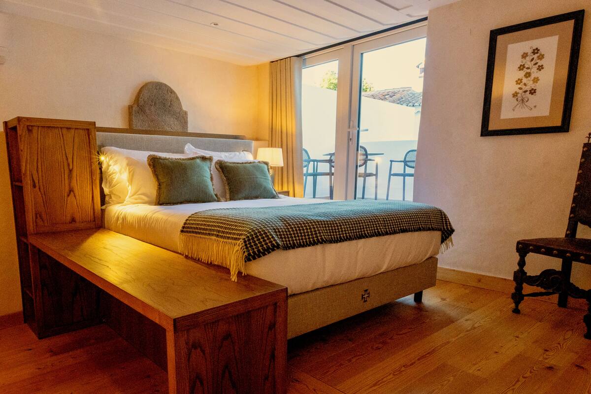 A bedroom with a wooden bed, a bench, a headboard, a bedside table, a lamp, a framed picture, and a view of the outside.