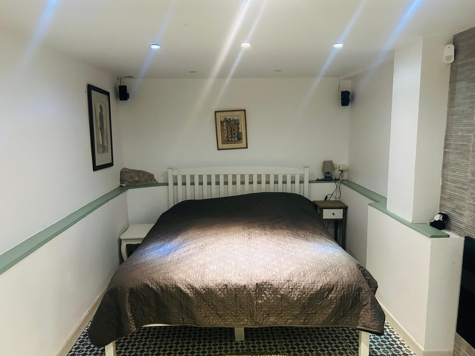 A room with a bed, a white radiator, a framed picture on the wall, and a small table with a lamp.