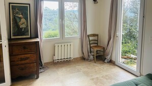 Ein Zimmer mit einem hölzernen Schminktisch, einem Sessel, einer Heizung und einem großen Fenster mit Vorhängen.