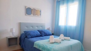 Een slaapkamer met een bed, kussens, een nachtkastje, een lamp, een raam met gordijnen en een decoratief kunstwerk.