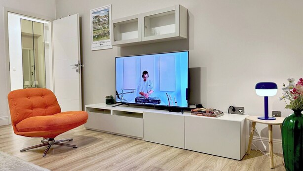 A modern living room with a flat-screen TV, a tufted orange chair, a white entertainment unit, and a green vase with flowers. - Vibehaus Center of Split City Only 400m From Beach (Split)