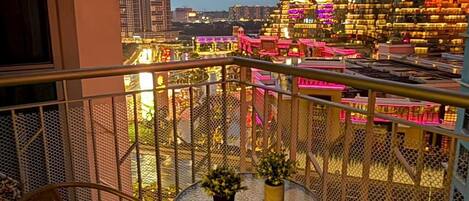 A balcony with a table and chairs, a view of a cityscape with illuminated buildings, and a mesh railing.