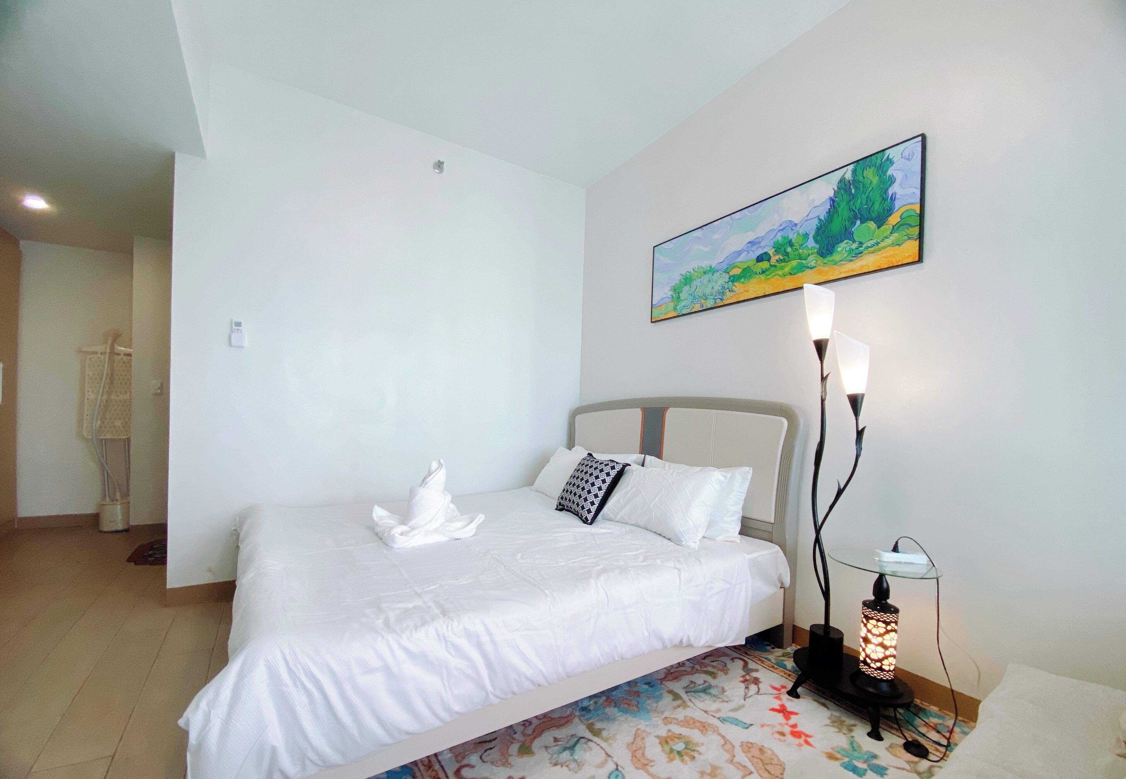 A neatly made bed with white linens and a decorative pillow, a painting on the wall, and a bedside table with a lamp.