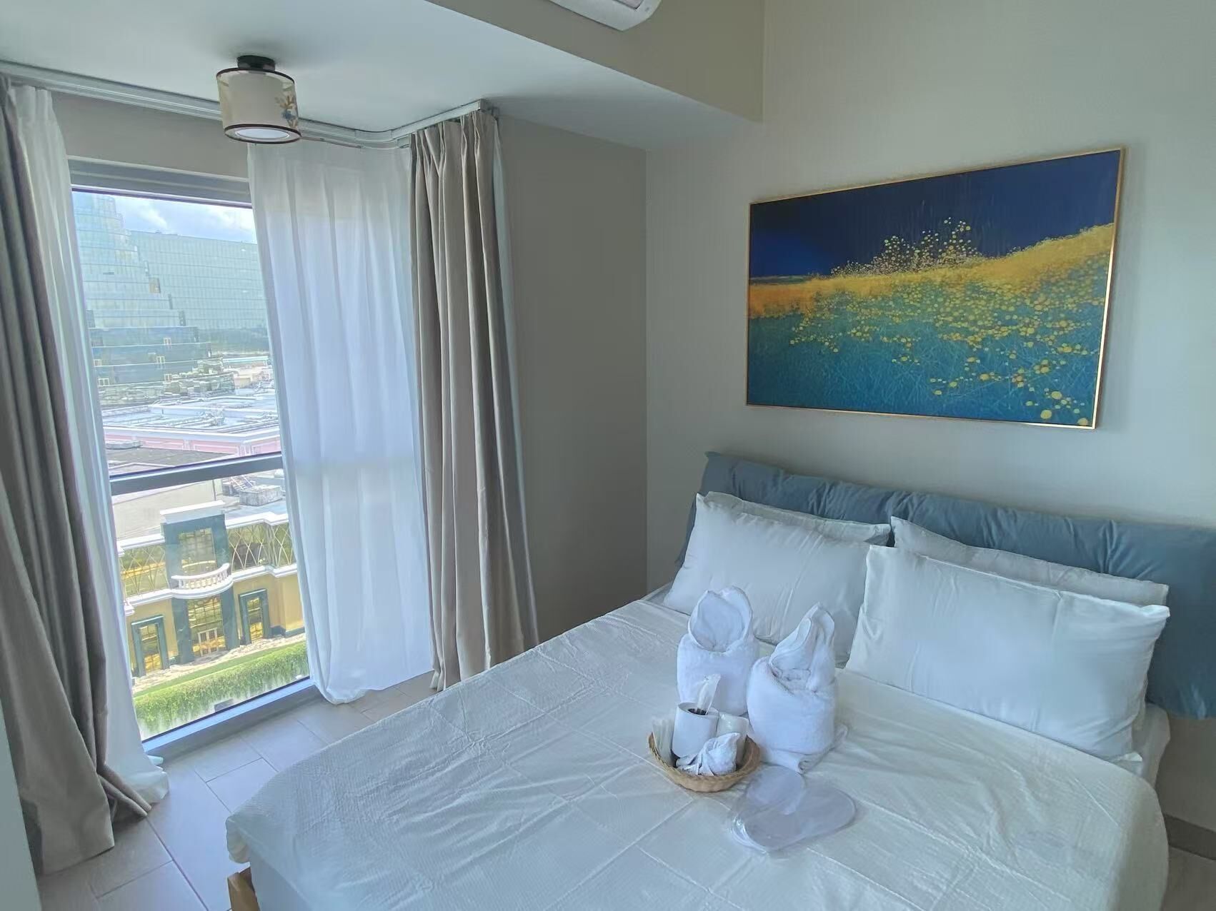 A hotel room with a large bed, a painting on the wall, and a view of buildings through the window.