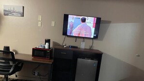 A hotel room with a television mounted on the wall, a desk with a microwave, a refrigerator, and a chair.