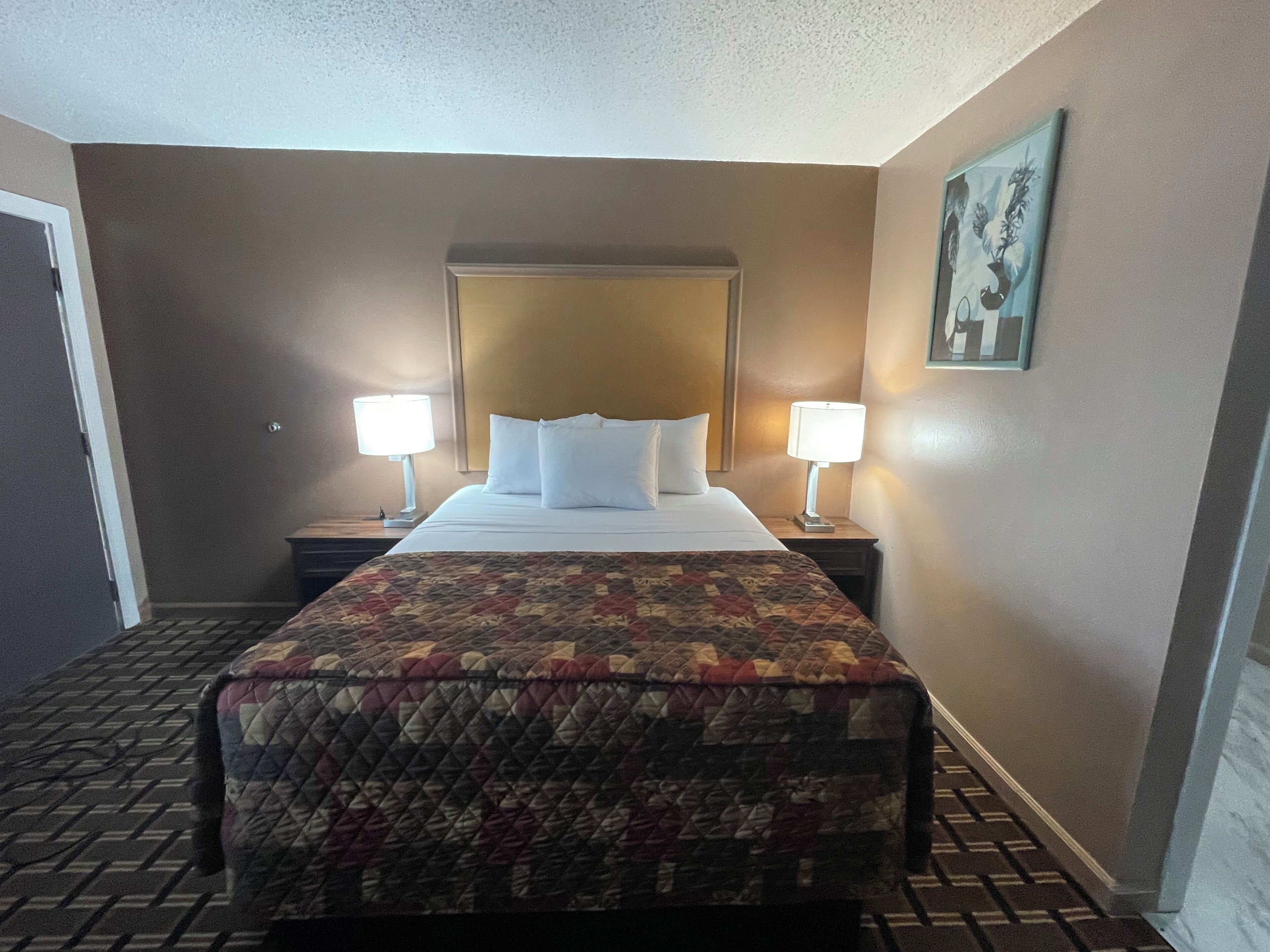 Una habitación de hotel con una cama, dos lámparas de noche, un cabecero, una colcha acolchada y un cuadro enmarcado en la pared.