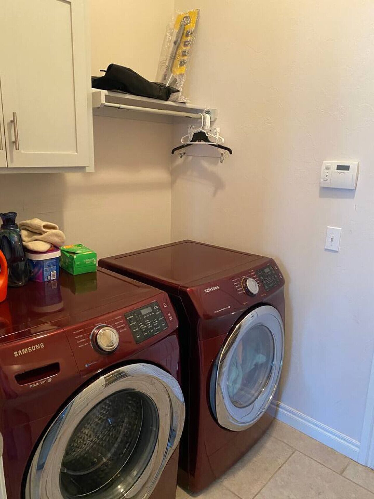 A laundry room with two Samsung washing machines, a shelf with a hanging rack, and a thermostat on the wall.