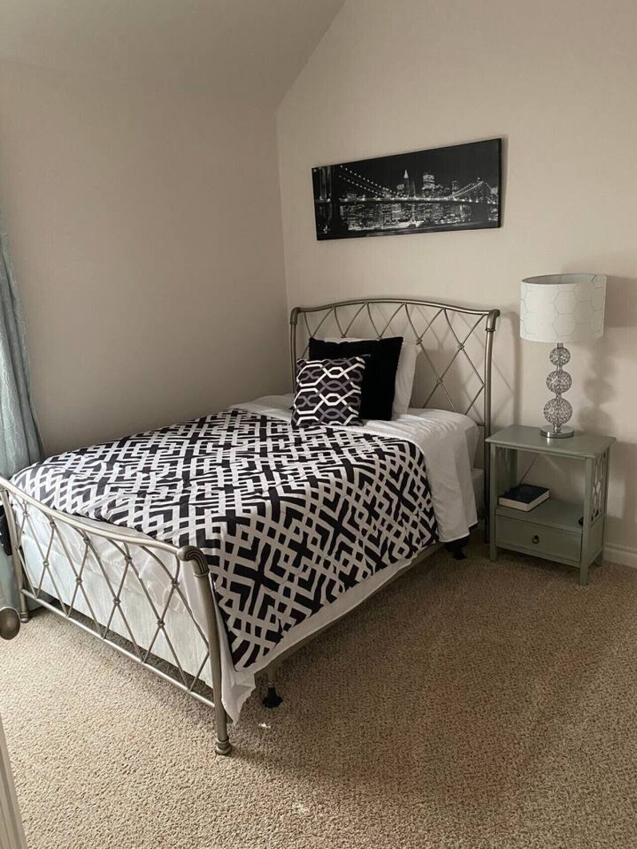 Un dormitorio ordenado con una cama de estructura metálica, una colcha estampada, una almohada y una lámpara de noche. En la pared, un cuadro con una vista urbana enmarcada.