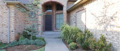 A brick house with a front door, a small front yard with shrubs, and a pathway leading to the door.