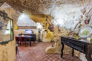 A rustic stone interior with a stone wall, a wooden table, chairs, a television, and a decorative plate. - La Casa di Simone (Palazzolo Acreide)