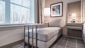 A hotel room with a bed, a bedside table, a chair, a window with curtains, a wall-mounted lamp, and a framed picture. - Hotel Bridgton (Bridgton)