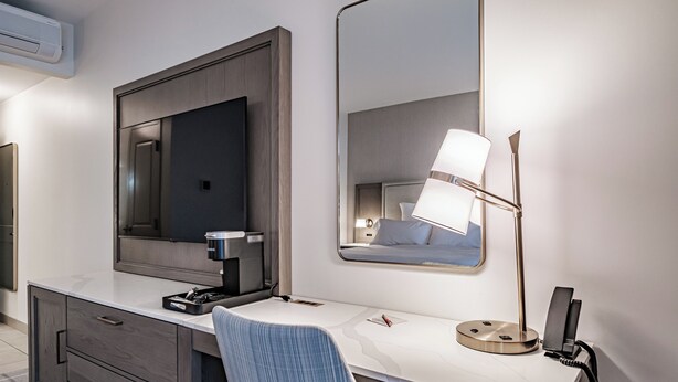 A modern hotel room with a large mirror, a desk with a lamp, a coffee maker, and a chair. - Hotel Bridgton (Bridgton)