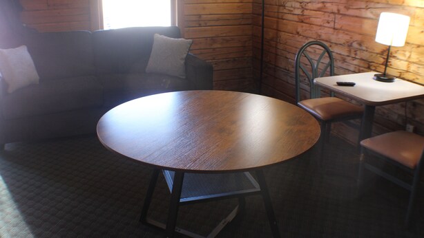 A room with a round wooden table, a green metal chair, a small table with a lamp, and a sofa with a pillow.