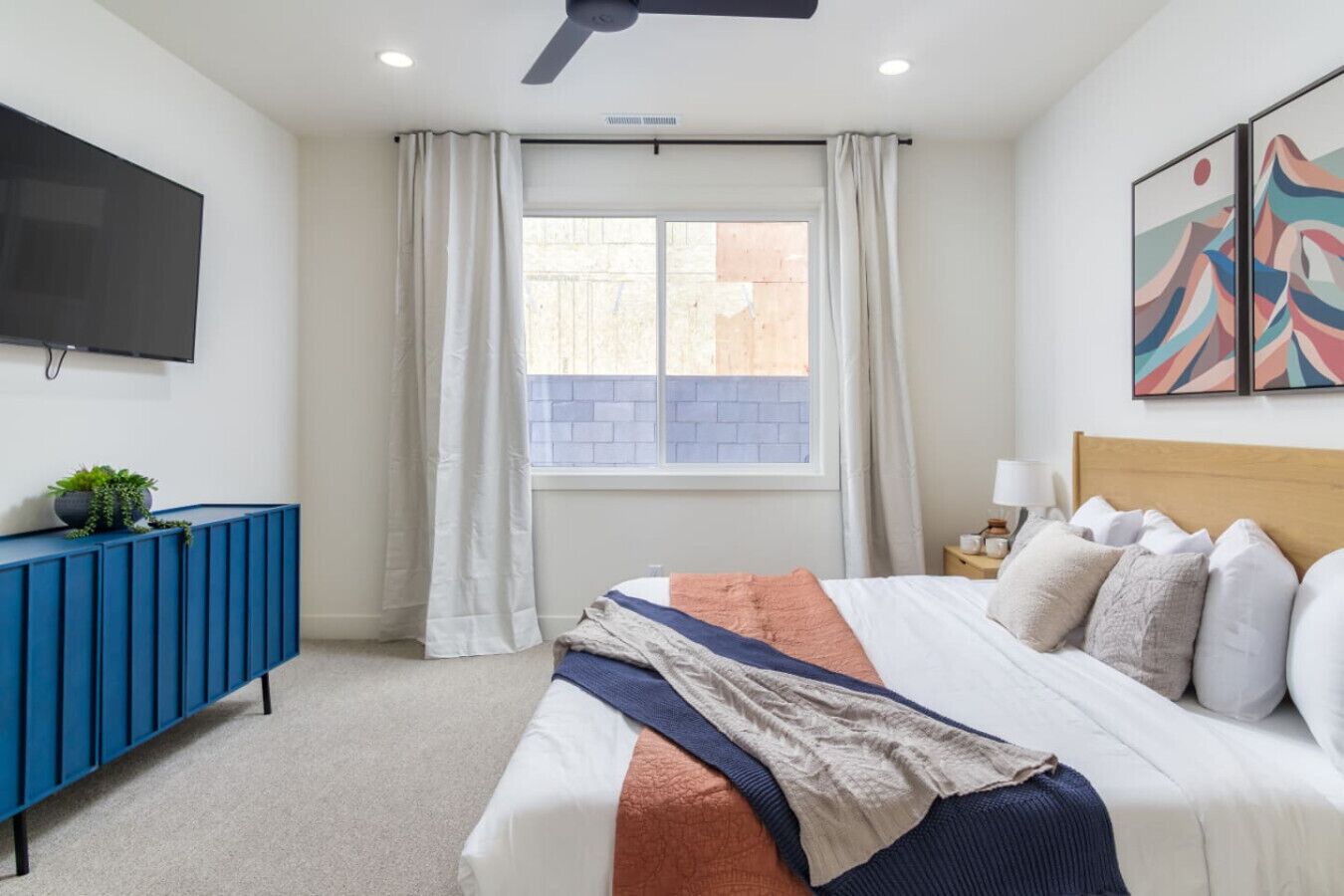 Une chambre à coucher avec un lit, une console bleue, un téléviseur, une fenêtre avec des rideaux et une œuvre d’art murale.