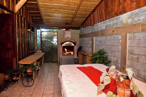 Tunnelmallinen huone, jossa on tiilestä muurattu takka, puuseinät, punaisella peitolla varustettu sänky ja puinen penkki.