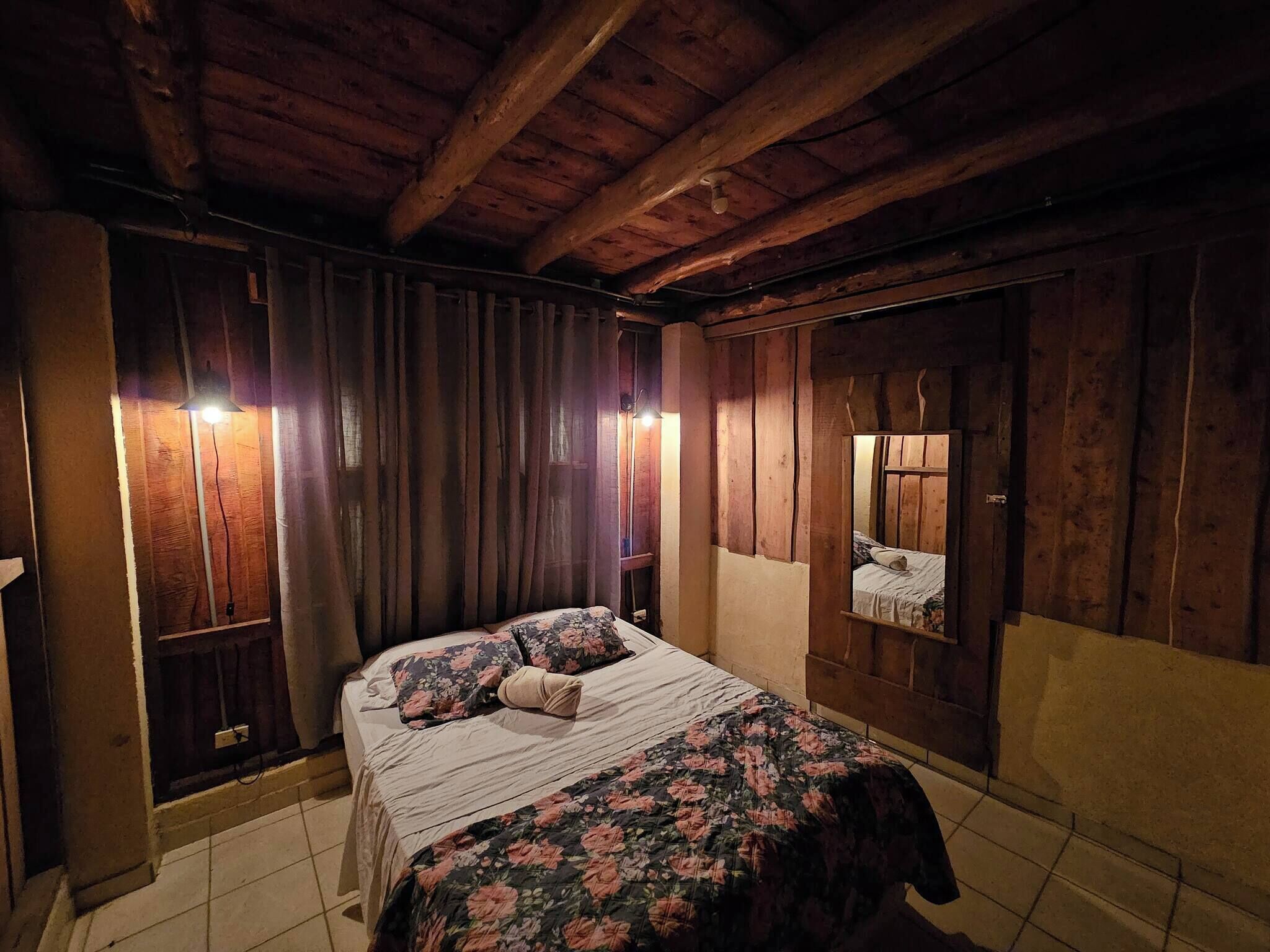 Una habitació amb bigues de fusta, un llit amb roba de llit floral, un mirall i cortines.