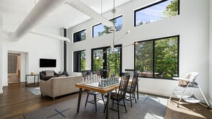 Un salón amplio con mesa de comedor, sillas, sofá y televisor. Ventanas grandes ofrecen luz natural y vistas al verdor.