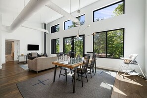 A spacious living room with a dining table, chairs, a sofa, and a TV. Large windows provide natural light and views of greenery. - Modern Mountain Retreat, Firepit, Grill and Fireplace (Old Fort)