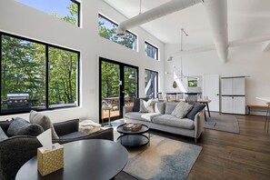 A spacious living room with large windows, a sofa, a coffee table, and a dining area with chairs. - Modern Mountain Retreat, Firepit, Grill and Fireplace (Old Fort)
