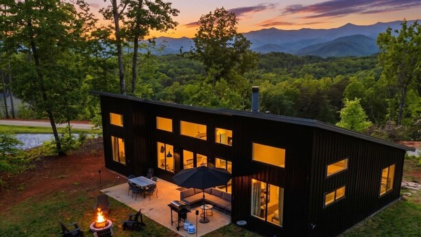 Una casa moderna de dos pisos con ventanas grandes, un patio con mobiliario exterior y un brasero.