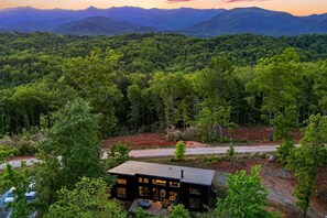 A modern house with a large deck, surrounded by dense forest and mountains in the background. - Modern Mountain Retreat, Firepit, Grill and Fireplace (Old Fort)